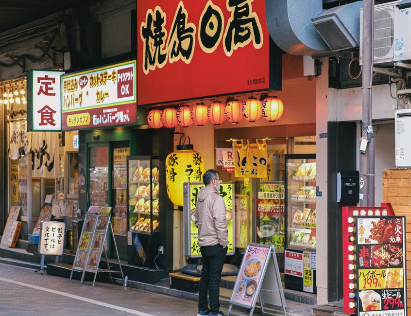 賑やかな通りにある日本食レストランの外に男性が立ち、メニューをじっくりと眺めている。明るい看板と日本語の文字が、居心地の良い雰囲気を醸し出している。