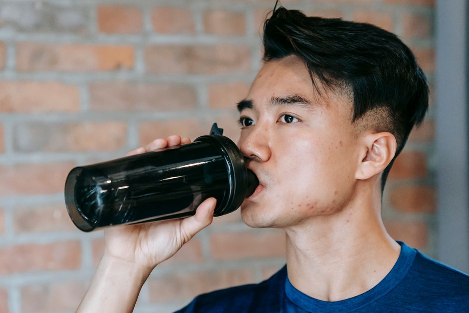 ジムの環境でシェイカーから水を飲む若い男性、集中力を保ちながら水分補給をしています。